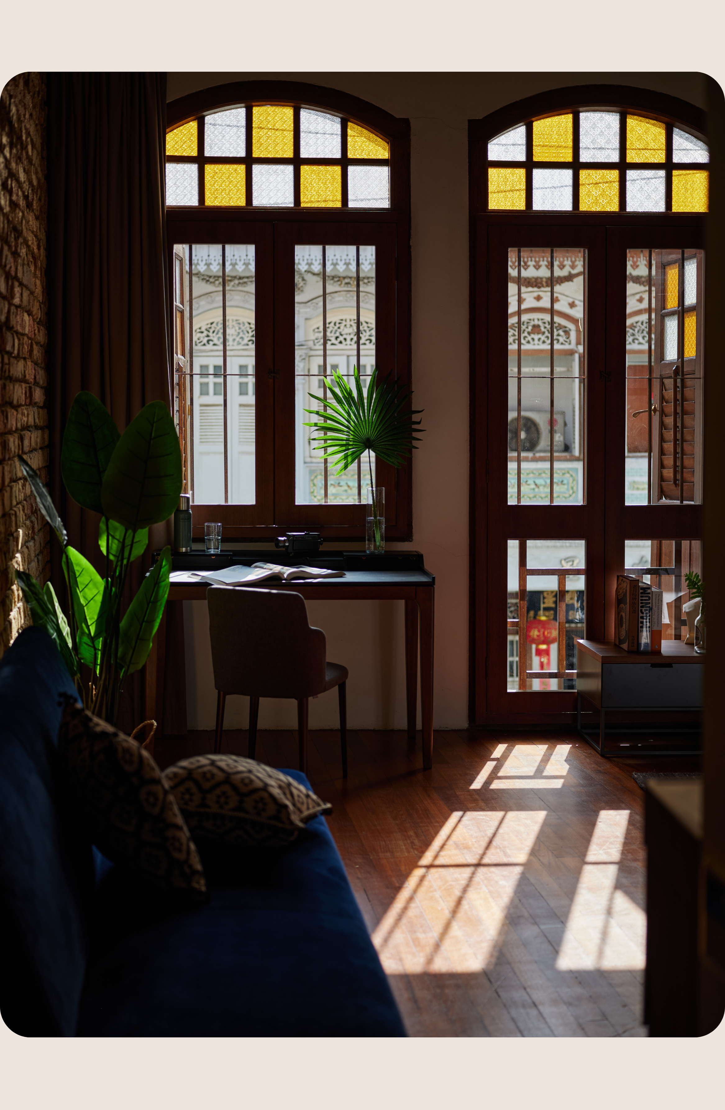 Sunlit interior with tall arched windows, stained glass panels, wooden floors, and a small desk beside indoor plants.