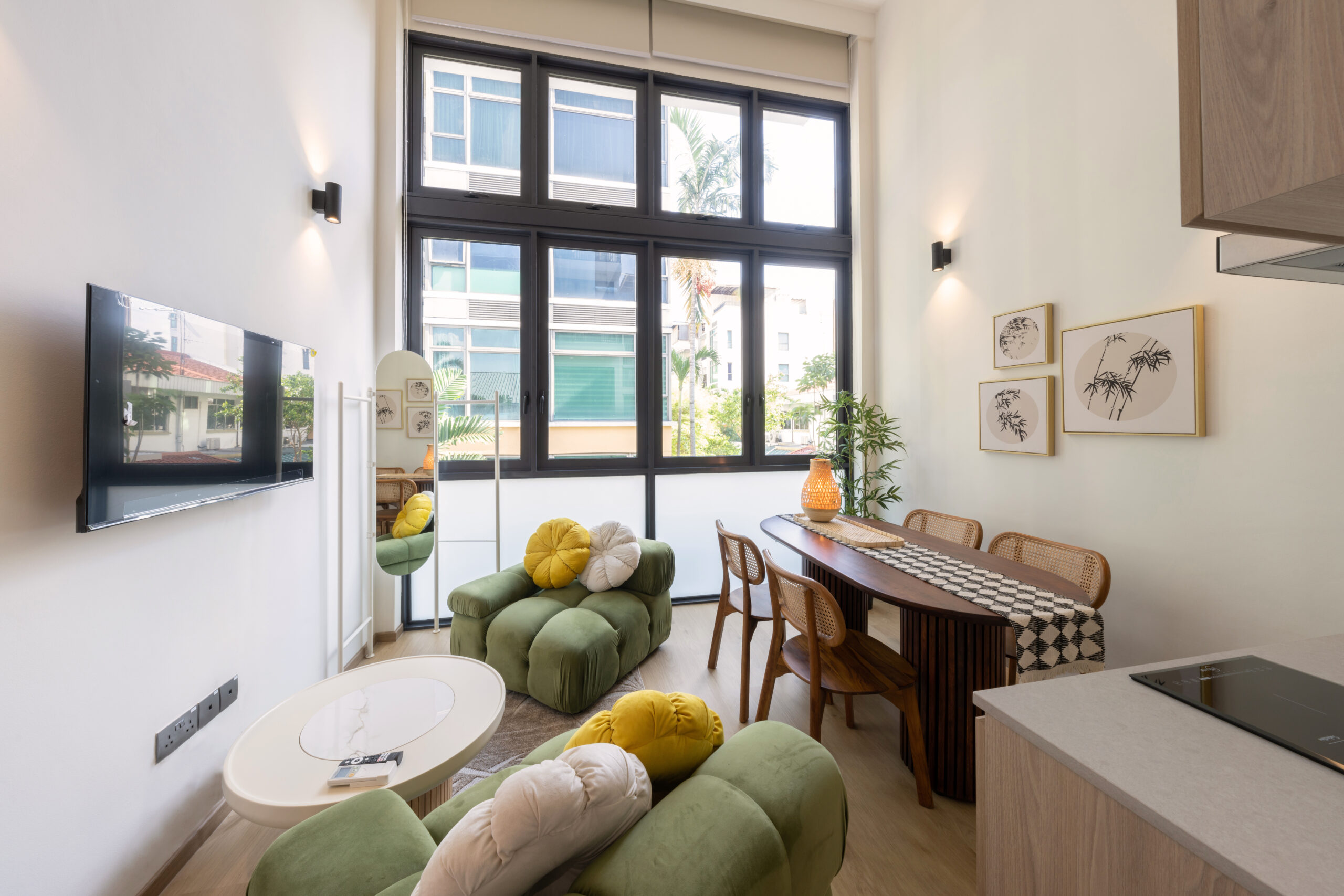 Bright, modern living and dining area with large black-framed windows, green upholstered lounge chairs, a wooden dining table with rattan chairs, wall-mounted TV, and neutral decor with indoor plants.