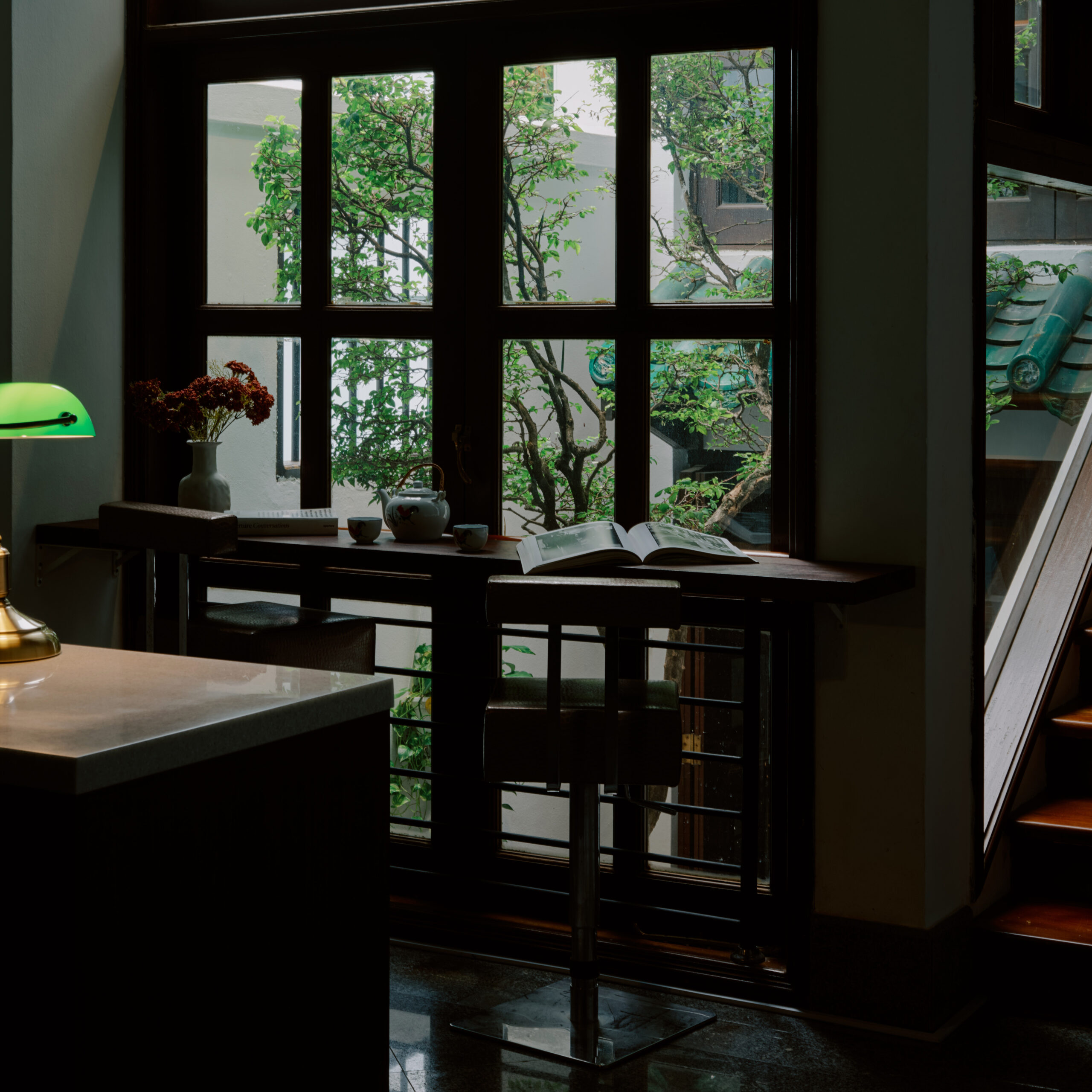 Window-side reading nook with a wooden counter, green desk lamp, and leafy garden view.